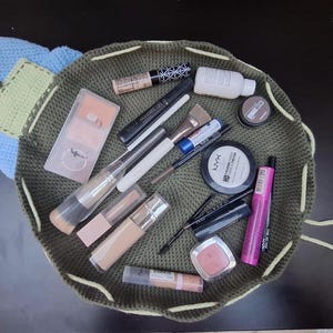 May include: A collection of makeup products, including brushes, mascara, foundation, and blush, are arranged inside a green crocheted drawstring bag. The bag is open, revealing the various cosmetic items. A blue and green crocheted flower is attached to the bag.