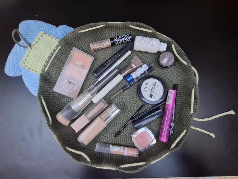 May include: A collection of makeup products, including brushes, mascara, foundation, and blush, are arranged inside a green crocheted drawstring bag. The bag is open, revealing the various cosmetic items. A blue and green crocheted flower is attached to the bag.
