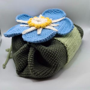 May include: A crocheted drawstring bag with a large blue flower on top. The flower has light blue petals, a yellow center, and white accents. The bag is primarily olive green with a light green section.