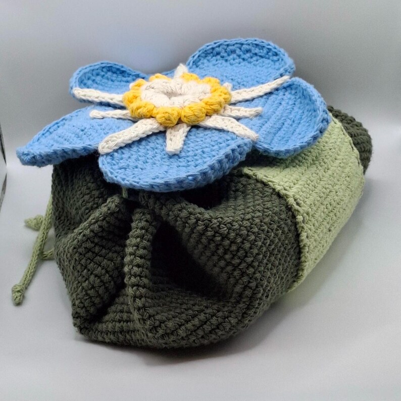 May include: A crocheted drawstring bag with a large blue flower on top. The flower has light blue petals, a yellow center, and white accents. The bag is primarily olive green with a light green section.