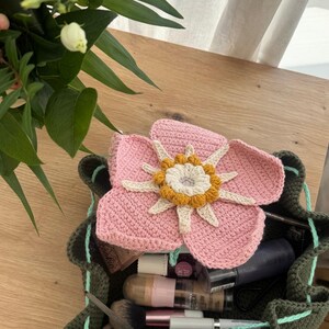 May include: A handmade, olive green crochet bag with a drawstring closure, filled with makeup items. A large, pink crochet flower with a yellow and white center rests on top. The bag is on a wooden surface.