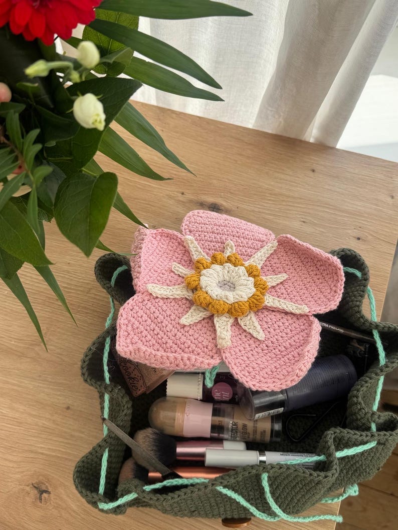 May include: A handmade, olive green crochet bag with a drawstring closure, filled with makeup items. A large, pink crochet flower with a yellow and white center rests on top. The bag is on a wooden surface.