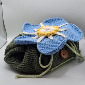 May include: A handmade crochet drawstring bag in olive green, topped with a large blue flower with yellow and white accents. The bag has a wooden button and green drawstring closure. The flower is the focal point of this unique, handcrafted bag.