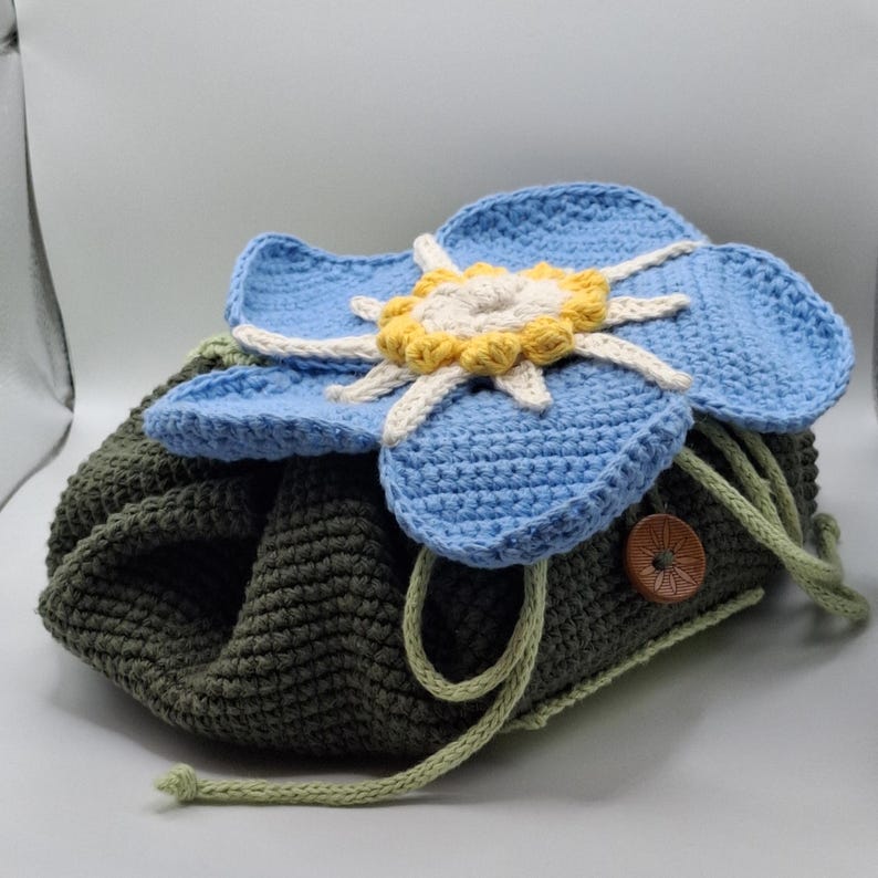May include: A handmade crochet drawstring bag in olive green, topped with a large blue flower with yellow and white accents. The bag has a wooden button and green drawstring closure. The flower is the focal point of this unique, handcrafted bag.