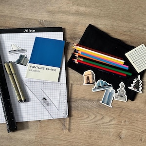 May include: A white gridded notebook with a black ruler, three black pens, a blue PANTONE colour swatch labelled "Skydiver 19-4151", and a black pencil case with coloured pencils and stickers of various buildings.