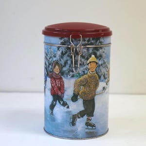 May include: A cylindrical metal storage canister with a deep red lid and a silver wire latch closure. The exterior features a winter scene illustration of two individuals ice skating on a frozen pond surrounded by snow-covered evergreen trees.