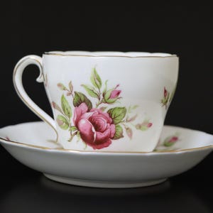 Peut inclure: Une tasse à thé et une soucoupe blanches avec un motif floral représentant des roses roses et des feuilles vertes. La tasse a un bord doré et la soucoupe a une bande dorée autour du bord.