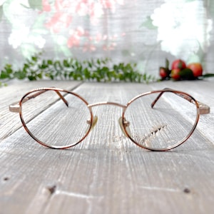 May include: A pair of gold-toned metal eyeglasses with round lenses. The glasses are sitting on a light wood surface.