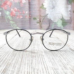 May include: Round eyeglasses with black frames and the text "MAINSTREET" on the right lens. The glasses have a silver bridge and nose pads. The background is a floral pattern on a light-colored surface.