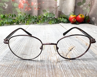 May include: A pair of burgundy-colored oval-shaped eyeglasses with a titanium frame. The glasses are on a wooden surface.