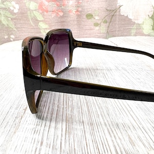 以下が含まれることがあります： 四角いフレームとグラデーションパープルのレンズが付いた、濃い茶色のサングラス。 サングラスは太いフレームとアームが付いています。 レンズは紫からクリアに変化します。 サングラスは視聴者の方を向いています。