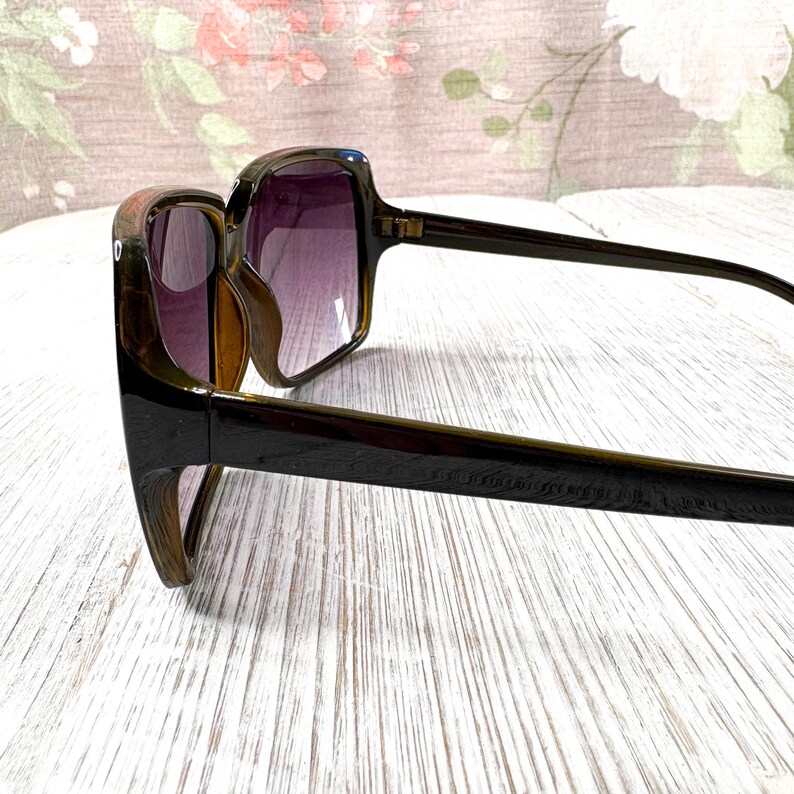 以下が含まれることがあります： 四角いフレームとグラデーションパープルのレンズが付いた、濃い茶色のサングラス。 サングラスは太いフレームとアームが付いています。 レンズは紫からクリアに変化します。 サングラスは視聴者の方を向いています。