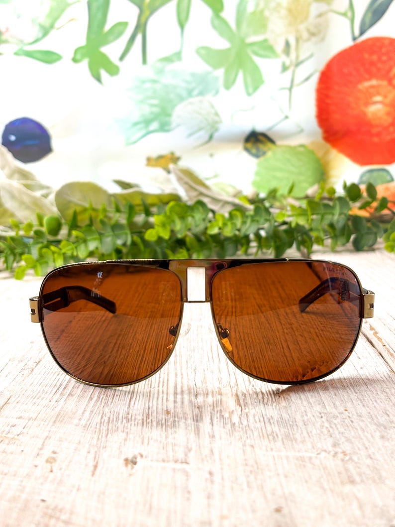 Op de afbeelding: Aviator zonnebril met een goudkleurig metalen montuur, een opvallende dubbele brug en amberkleurige glazen. De pootjes zijn dun en van metaal, verbonden met het voorste montuur via zichtbare scharnieren, wat zorgt voor een klassieke uitstraling.