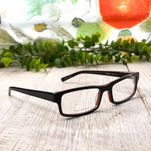 May include: Rectangular eyeglasses with a dark brown frame and clear demo lenses. The glasses have a subtle red interior and are set against a backdrop of a floral pattern and greenery. The text "DEMO LENS ONLY" is visible on the lens.