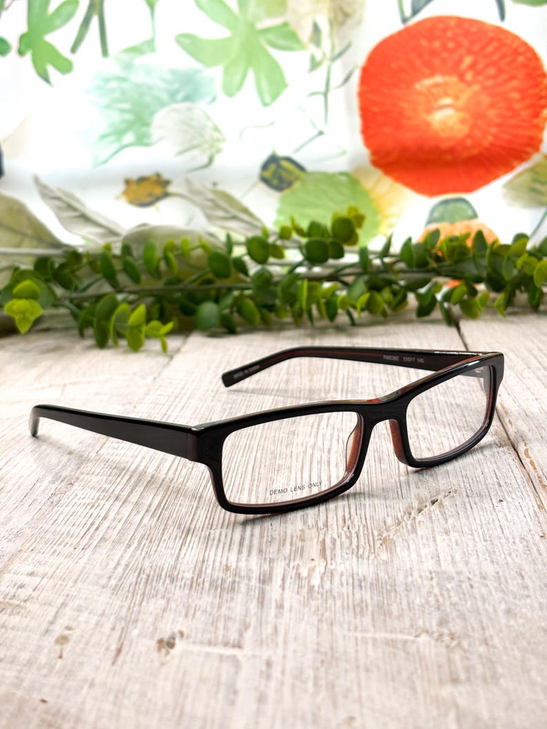 Pode incluir: &Oacute;culos retangulares castanhos escuros com lentes de demonstra&ccedil;&atilde;o transparentes. Os &oacute;culos t&ecirc;m um interior vermelho subtil e est&atilde;o sobre um fundo de padr&otilde;es florais e vegeta&ccedil;&atilde;o. O texto "DEMO LENS ONLY" &eacute; vis&iacute;vel na lente.