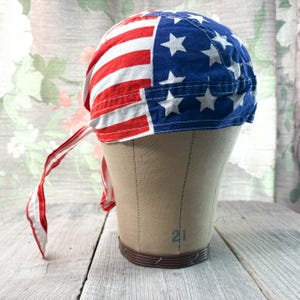 May include: A patriotic bandana featuring the American flag design. The bandana is divided into two sections: one with red and white stripes, and the other with a blue field and white stars. The bandana has ties on the back.