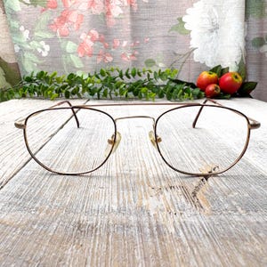 May include: A pair of vintage eyeglasses with a gold-colored metal frame and clear lenses. The glasses have a double bridge design and are resting on a weathered, light-colored wooden surface. Decorative greenery and small red apples are in the background.