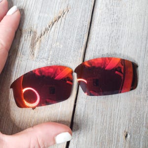 Puede incluir: Dos lentes de repuesto de color rojo espejo para gafas de sol. Las lentes son curvadas y tienen una superficie reflectante.