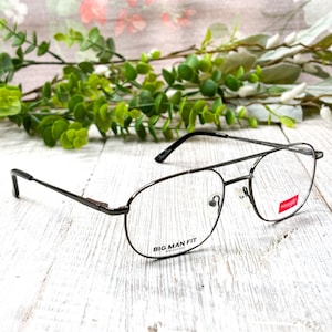 Puede incluir: Un par de gafas con montura de metal oscuro. Las gafas tienen doble puente y forma rectangular. El texto "BIG MAN FIT" está impreso en la montura. La marca "Wrangler" es visible en la lente.