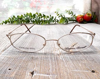 May include: Gold-tone metal eyeglasses with a thin frame and oval lenses. The frame is inscribed with "Lauren Bacall" and "Stainless Steel".