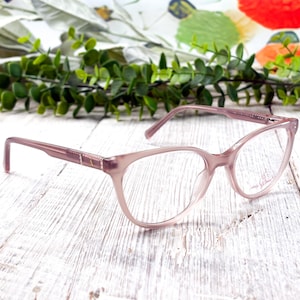 May include: Pale pink cat-eye spectacles with clear lenses. The frames have a subtle sparkle detail near the temples. The glasses rest on a white, weathered wooden surface, with a floral background.