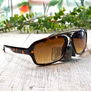 May include: Tortoiseshell sunglasses with gradient brown lenses. The sunglasses have a rectangular frame and a tag that reads "FASHION UV 400". The sunglasses are on a light wood surface.