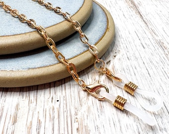 May include: Gold-colored eyeglass chain with a delicate chain design. The chain has a lobster clasp closure and clear rubber loops to attach to glasses. The chain is displayed on a light blue ceramic plate and a white wooden surface.