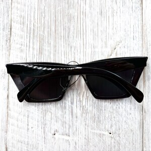 Puede incluir: Gafas de sol negras con un dise&ntilde;o elegante y angular. Las monturas son brillantes y rectangulares, con una forma ligeramente sobredimensionada. Las lentes son oscuras, proporcionando una cobertura total. Las gafas se muestran sobre un fondo de madera blanca.