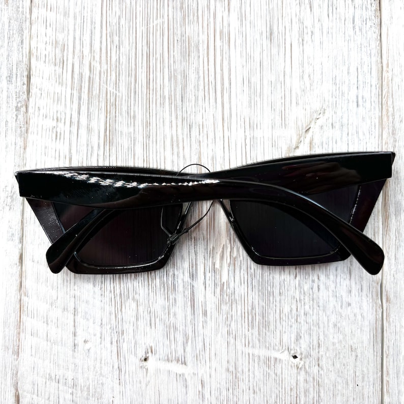 Puede incluir: Gafas de sol negras con un dise&ntilde;o elegante y angular. Las monturas son brillantes y rectangulares, con una forma ligeramente sobredimensionada. Las lentes son oscuras, proporcionando una cobertura total. Las gafas se muestran sobre un fondo de madera blanca.