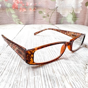 Pode incluir: Um par de óculos com armação e hastes com padrão tartaruga. As lentes são transparentes e as armações são de cor castanha quente. Os óculos estão sobre uma superfície de madeira branca.