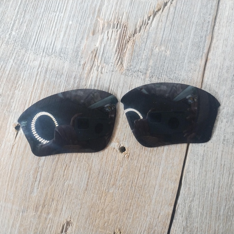 May include: Two black replacement lenses for sunglasses. The lenses are curved and have a dark tint.