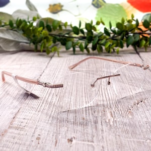 May include: Rimless eyeglasses with a light pink frame and clear lenses. The temples are adorned with small, sparkling embellishments. The glasses rest on a weathered, light-colored wooden surface, with greenery in the background.