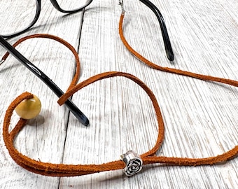 May include: A brown leather eyeglass chain with a silver rose accent and a small, round, light-colored bead. Black eyeglasses are visible in the background, resting on a white wooden surface. The chain is designed to hold glasses.