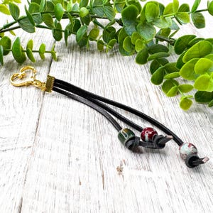 May include: A black leather keychain with a gold-colored heart-shaped clasp. The keychain features three black leather strands, each adorned with a decorative bead in shades of green, red, and white. The background is a white wooden surface.