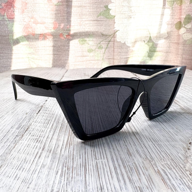 Puede incluir: Gafas de sol negras con un dise&ntilde;o geom&eacute;trico &uacute;nico. Las monturas son brillantes y angulares, con una forma afilada y puntiaguda. Las lentes son oscuras y las gafas de sol tienen una est&eacute;tica moderna y atrevida. El texto "CHINA NFASH1" est&aacute; impreso en la montura.
