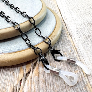 Peut inclure: Chaîne de lunettes en métal noir avec un motif de chaîne à maillons. La chaîne a des fermoirs noirs et des boucles en caoutchouc transparentes pour la fixation des lunettes. La chaîne repose sur une assiette en céramique bleu clair et beige.