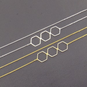 Op de afbeelding: Twee kettingen met drie zeshoekige hangerbedels. De bovenste ketting is zilver en de onderste ketting is goud.