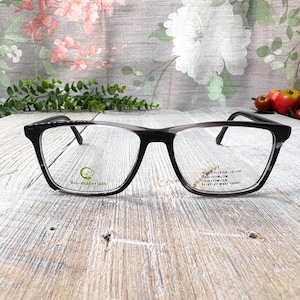 Puede incluir: Gafas rectangulares negras y grises con lentes transparentes. Las gafas tienen un logotipo de biomateriales en la lente izquierda y texto en la lente derecha. Las gafas están sobre una superficie de madera clara.