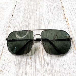 Pode incluir: &Oacute;culos de sol aviador com arma&ccedil;&atilde;o prateada e lentes verde escuro. Os &oacute;culos t&ecirc;m uma ponte dupla e uma forma retangular cl&aacute;ssica. As lentes s&atilde;o ligeiramente refletivas e a arma&ccedil;&atilde;o &eacute; prateada brilhante. Sobre uma superf&iacute;cie de madeira branca.