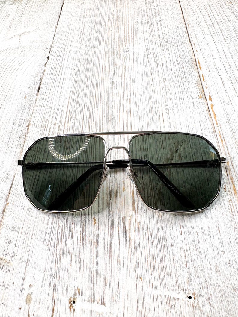 Pode incluir: &Oacute;culos de sol aviador com arma&ccedil;&atilde;o prateada e lentes verde escuro. Os &oacute;culos t&ecirc;m uma ponte dupla e uma forma retangular cl&aacute;ssica. As lentes s&atilde;o ligeiramente refletivas e a arma&ccedil;&atilde;o &eacute; prateada brilhante. Sobre uma superf&iacute;cie de madeira branca.