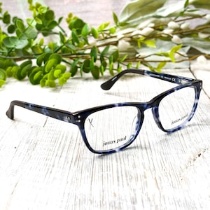 May include: Eyeglasses with a blue and white marbled pattern on the frames. The arms are black. The brand name "jonas paul" is printed on the lower right lens. The glasses are set against a white wooden surface with greenery in the background.