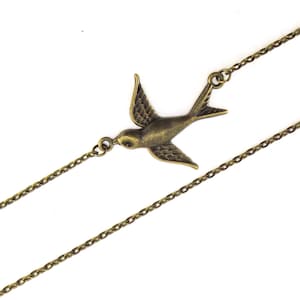 Puede incluir: Un collar dorado con una cadena delicada y un colgante de pájaro. El pájaro está en vuelo con las alas extendidas.