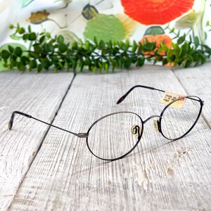 May include: Oval-shaped eyeglasses with thin, dark metal frames and gold-toned nose pads. A small beige paper tag is attached to the right temple, featuring the text SAKURA 185, 51 18 140, and AMBER. The temples have dark brown, mottled tips.
