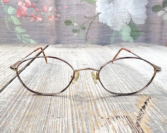 May include: Oval-shaped eyeglasses with a thin, bronze-colored metal frame. The lenses are clear, and the nose pads are a light beige. The glasses are set against a backdrop of a floral pattern on a wooden surface.