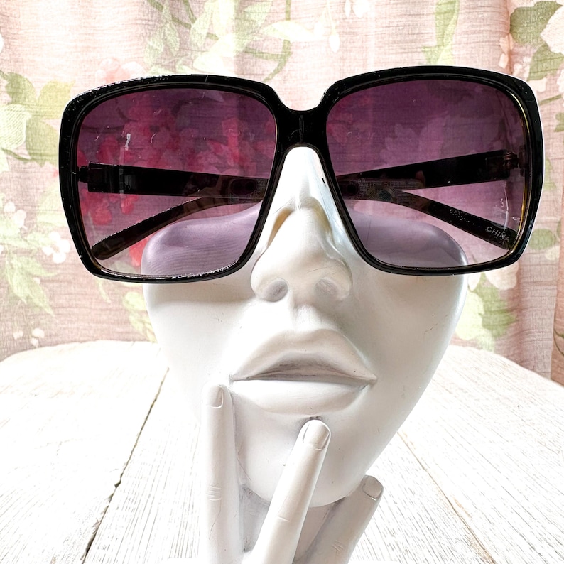 以下が含まれることがあります： 紫色のグラデーションレンズが付いた黒いスクエアフレームのサングラス。 サングラスは太いフレームで、白いマネキンの頭に表示されています。 レンズは上部で濃い色合いから下部で明るい色合いに変化します。