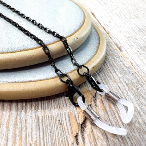 May include: Black metal eyeglasses chain with a rectangular link design. The chain has black clasps and white rubber loops to attach to the glasses. The chain is displayed on a light blue and tan ceramic plate.