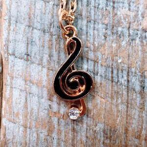 Puede incluir: Collar dorado con un colgante de clave de sol negro y una pequeña gema transparente. El colgante cuelga de una fina cadena dorada. Fondo de madera.