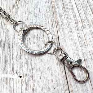 May include: Silver-toned necklace with a large hammered-texture circle pendant and a lobster clasp. The necklace has a double chain and is designed for attaching keys or other small items. The background is a light-colored wooden surface.