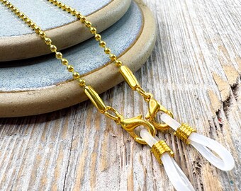 May include: Gold-colored eyeglass chain with a delicate ball chain design. The chain features gold clasps and clear, teardrop-shaped attachments for the glasses. The chain rests on a light blue ceramic plate and a wooden surface.