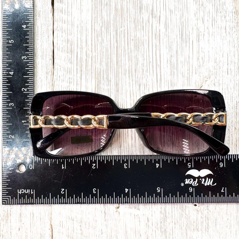Puede incluir: Gafas de sol negras con detalle de cadena dorada en la montura. Las gafas de sol miden aproximadamente 15 cm de ancho. Las lentes son de color degradado, pasando de oscuro a claro. Las gafas est&aacute;n sobre una superficie de madera blanca.